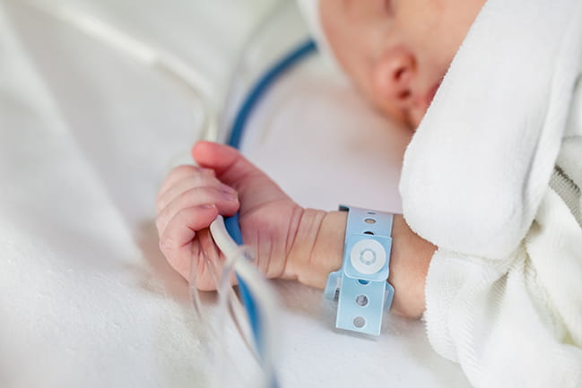 Caucasian infant in hospital bed clutching hoses and cords and wearing hospital identification band.